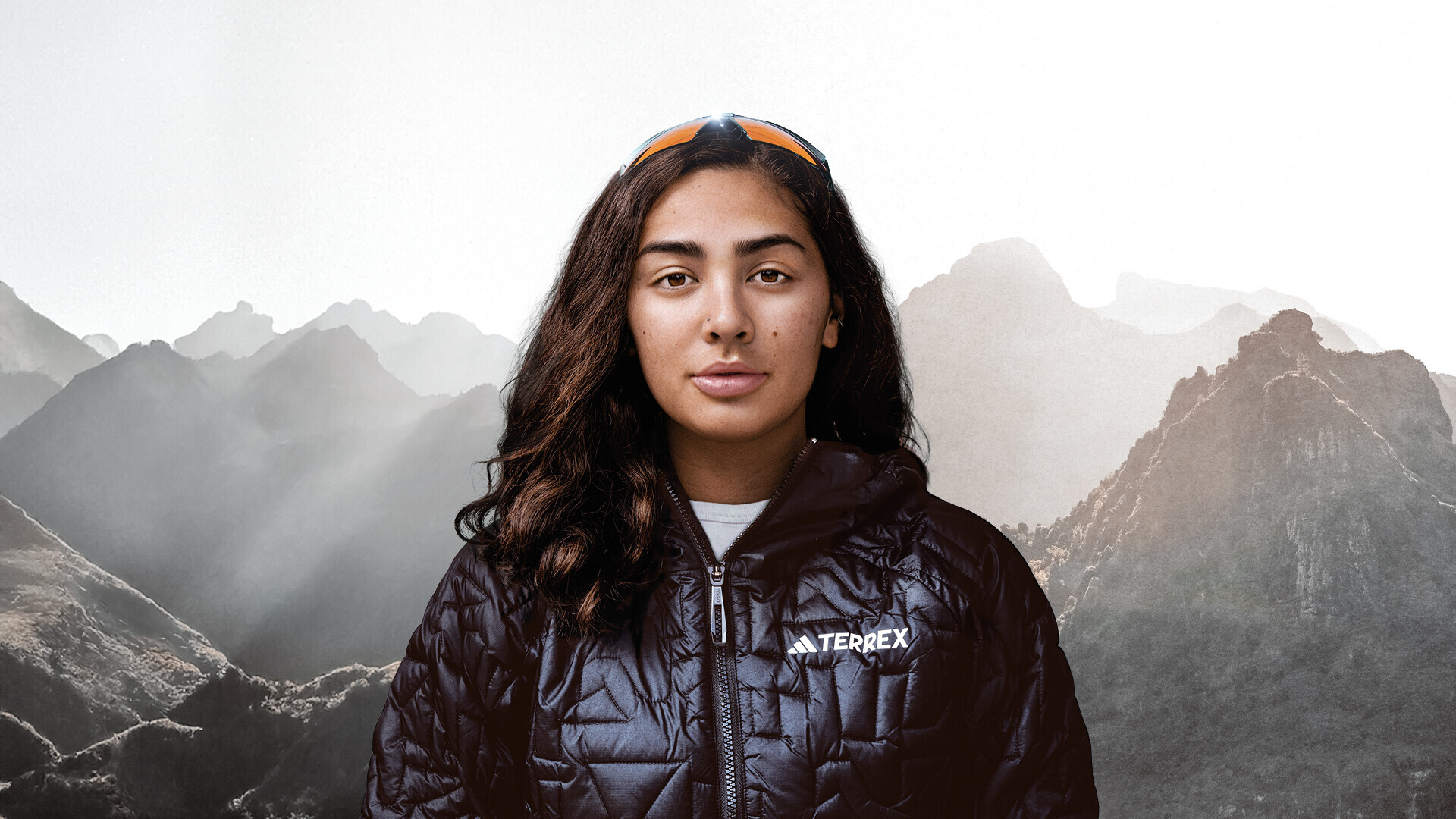 Portrait de Samantha Soriano regardant directement la cam&eacute;ra. Elle porte une veste d'ext&eacute;rieur noire et des lunettes de soleil sur la t&ecirc;te. En arri&egrave;re-plan, on voit des sommets de montagnes embrum&eacute;s.