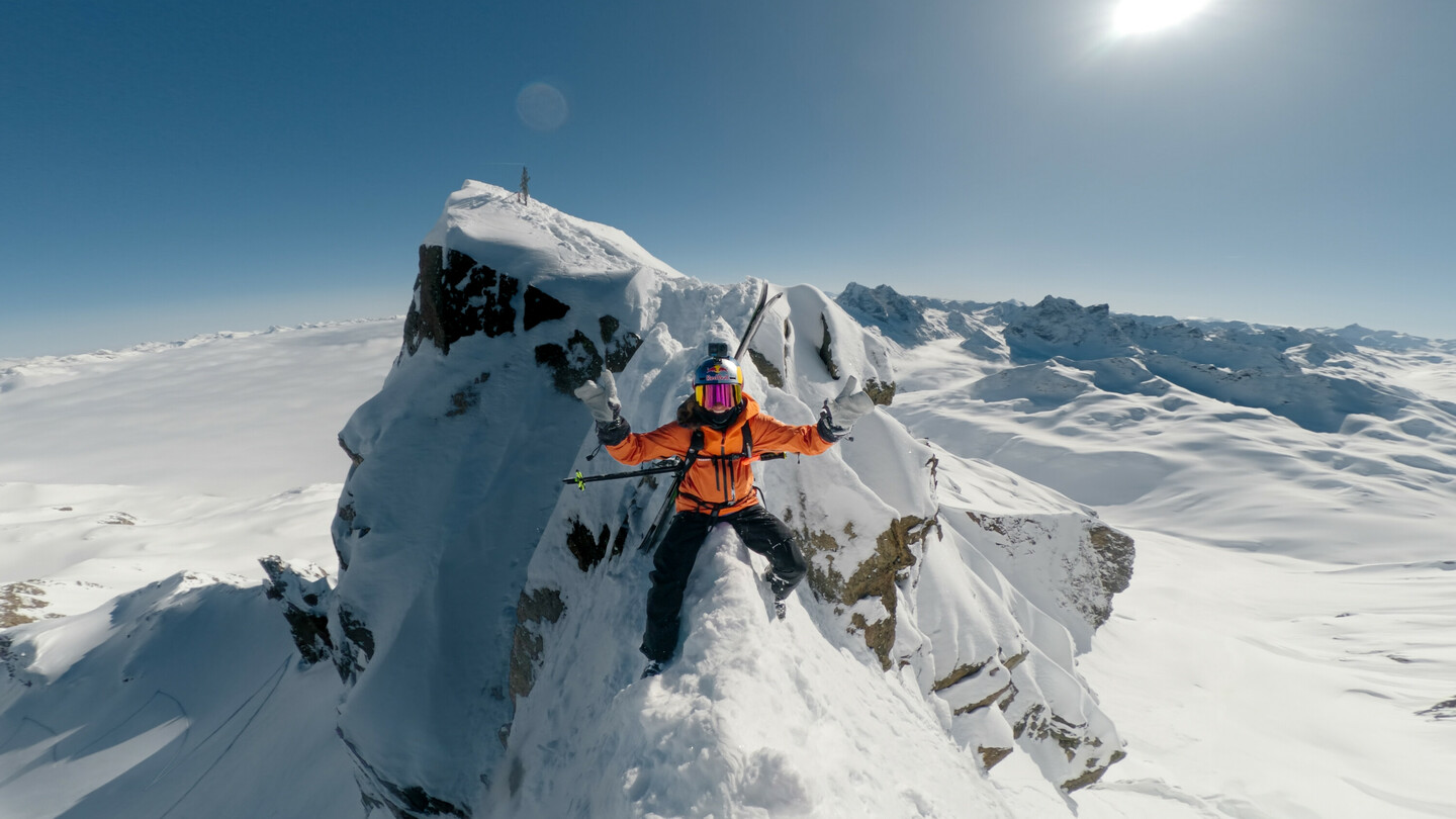 Skieur en veste orange en équilibre sur une étroite corniche enneigée, entouré d'un paysage hivernal spectaculaire sous un ciel lumineux.