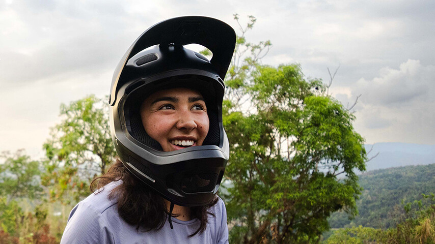 Samantha Soriano souriante avec un casque intégral noir en plein air. À l'arrière-plan, on voit des arbres et une région vallonnée boisée, avec un ciel légèrement nuageux.