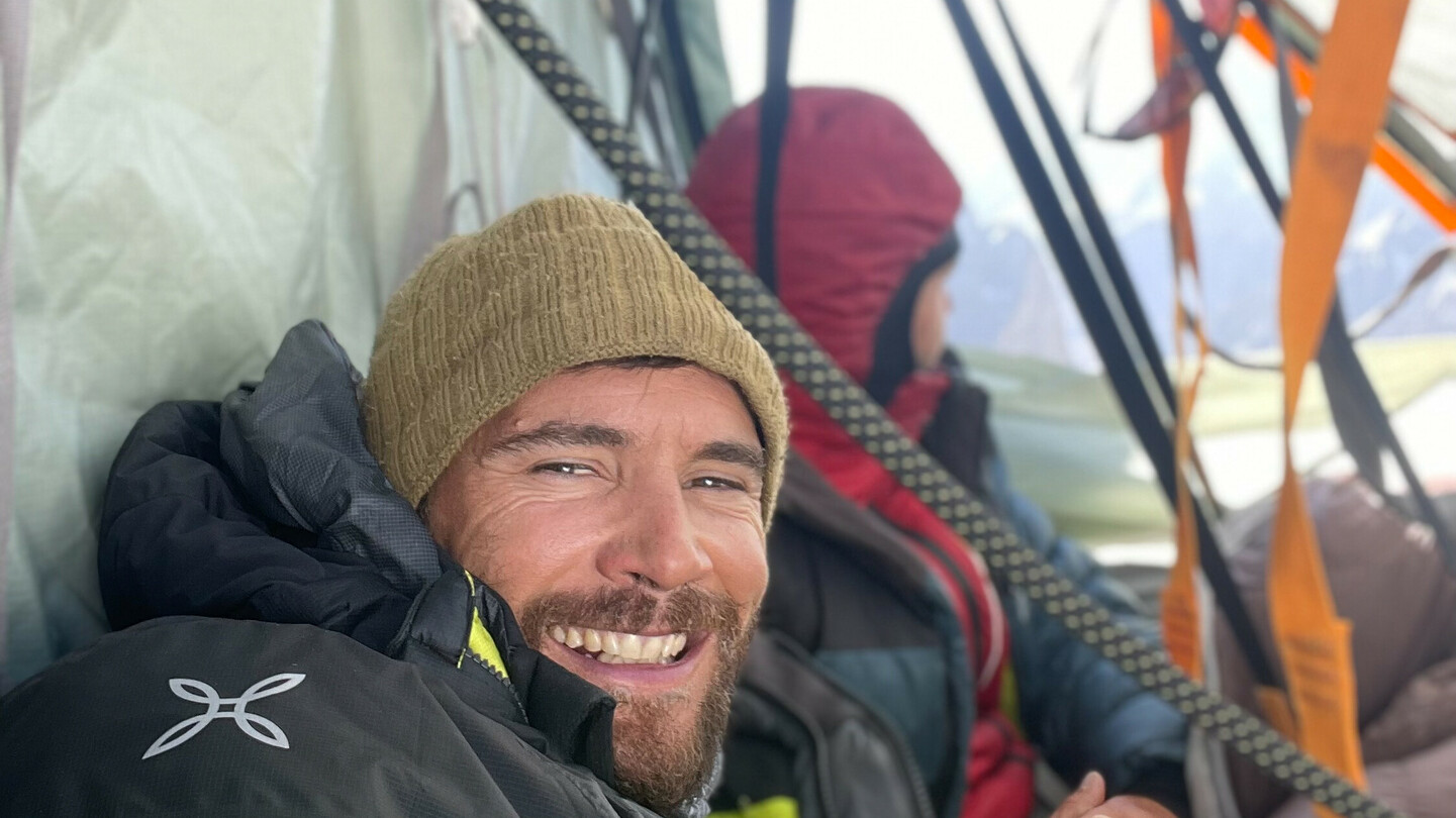 Eduard Marín smiling inside a tent during a mountain expedition, wrapped in warm outdoor clothing, with another climber in the background.