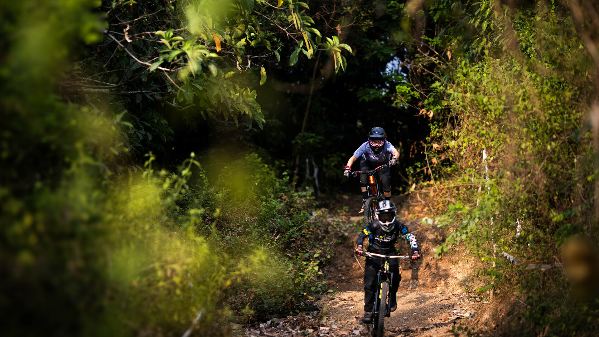 Deux vététistes descendent un sentier étroit bordé d'arbres. Le cycliste de devant porte un équipement noir avec un casque, tandis que celui de derrière est habillé en gris avec des lunettes de protection.