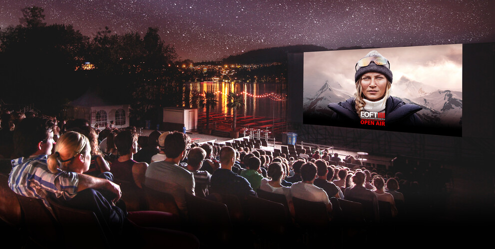 Un cinéma en plein air au bord du lac avec un large public regardant un film sous le ciel étoilé. Sur l'écran, une femme en tenue d'extérieur avec des montagnes en arrière-plan, accompagné du texte “EOFT OPEN AIR”.