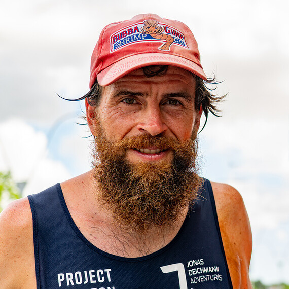 Portrait de Jonas Deichmann, un homme avec une barbe &eacute;paisse et une casquette rouge arborant le logo Bubba Gump Shrimp Co. Il porte un d&eacute;bardeur fonc&eacute;.